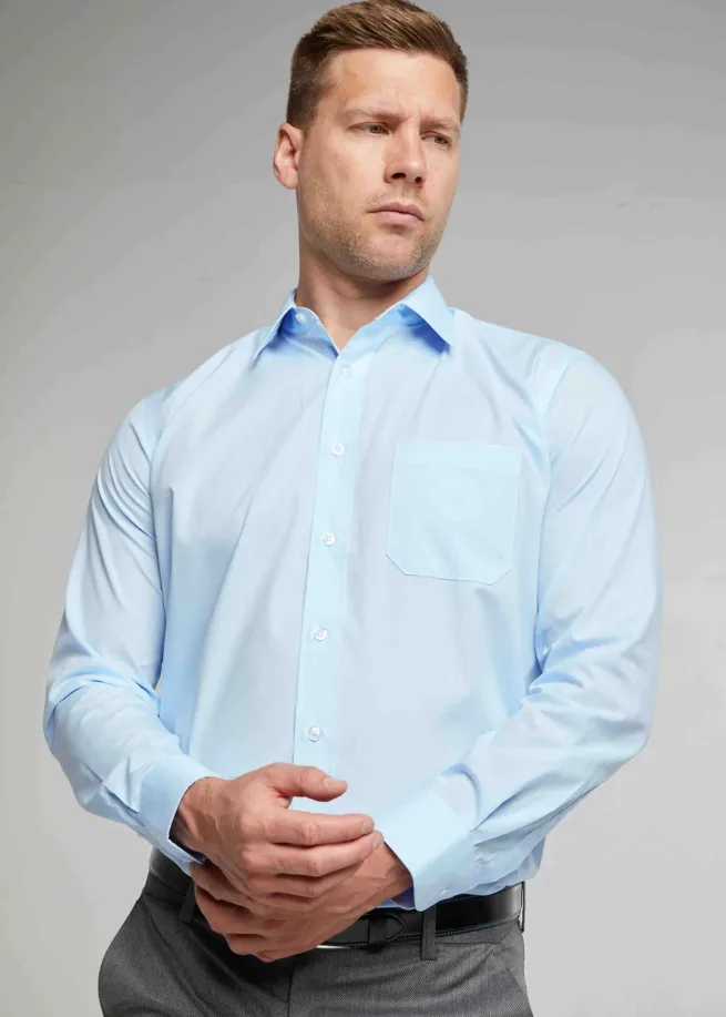 Man standing wearing light blue long sleeve shirt. One hand is clasping the other cuff.
