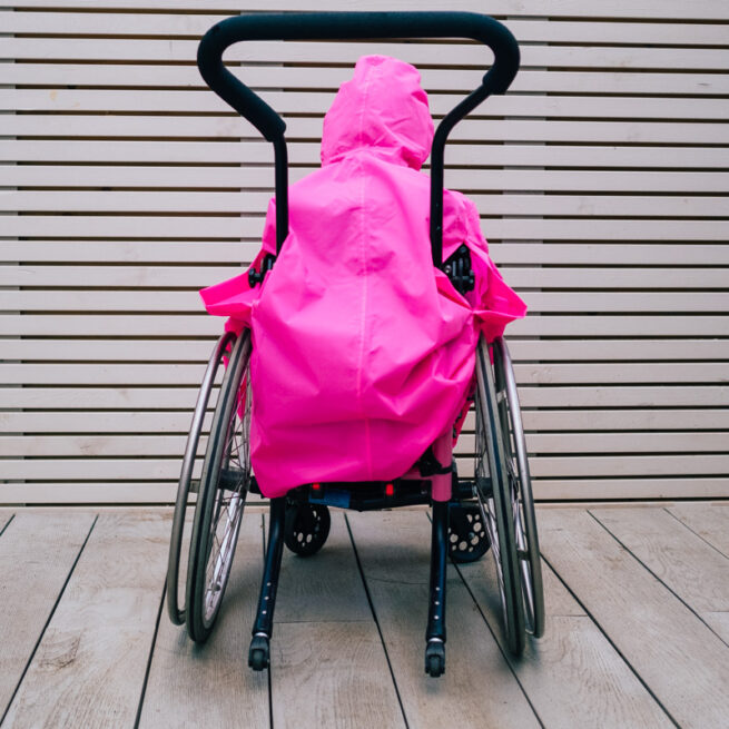 Back view of girl in wheelchair wearing pink waterproof coverall with hood up